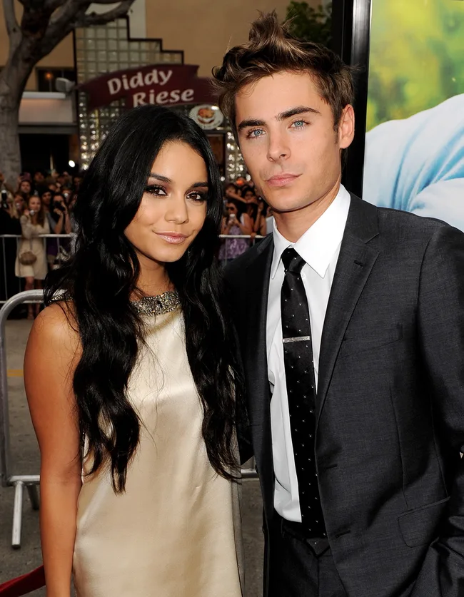 A woman in a light dress and a man in a suit stand together at an event with a crowd in the background.