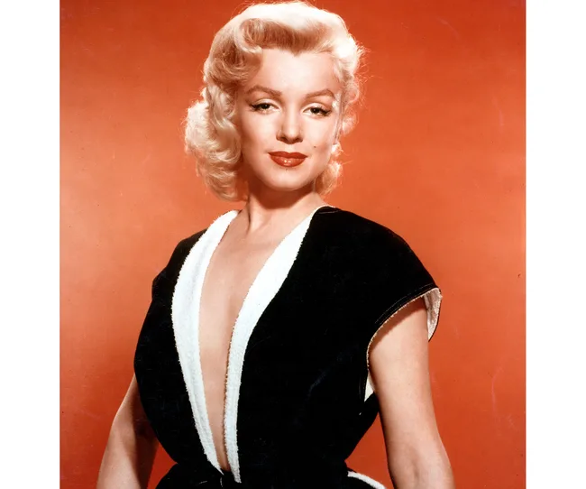 Blonde woman in black and white outfit with a plunging neckline, posing against an orange background.