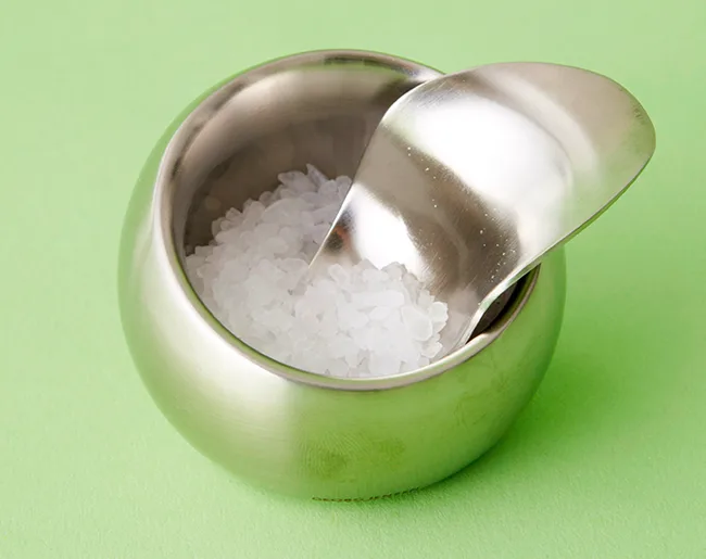 Stainless steel bowl with sea salt and a small scoop on a light green surface.