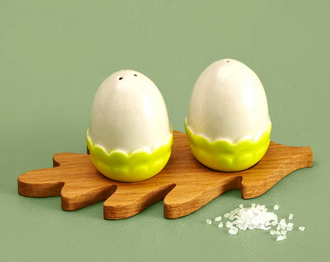 Ceramic egg-shaped salt and pepper shakers with a green base on a wooden leaf tray, with spilled salt beside.