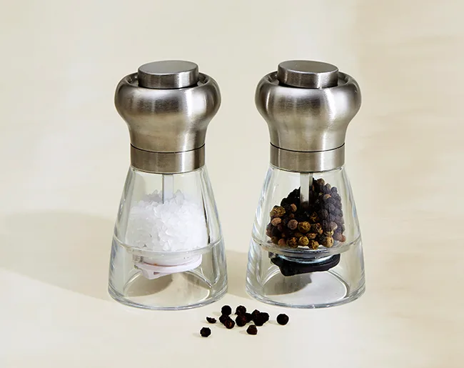 Salt and pepper grinders with stainless steel tops on a light background. Black peppercorns scattered in front.