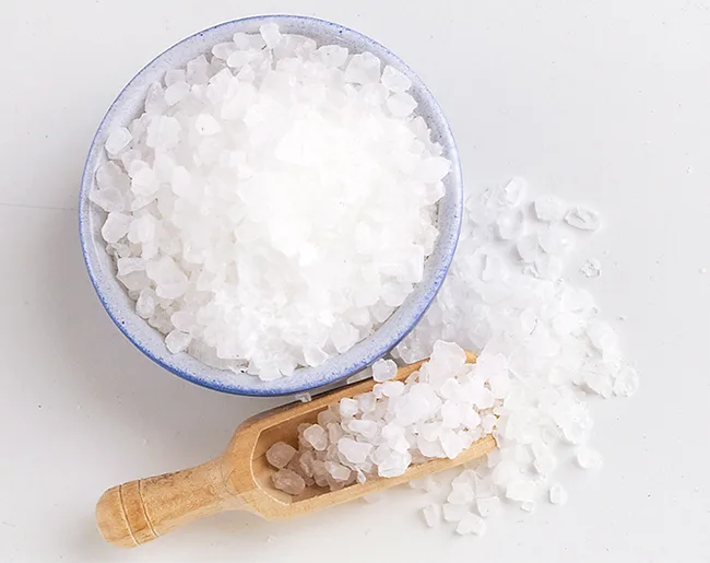 Bowl of coarse sea salt with a wooden scoop on a white surface.