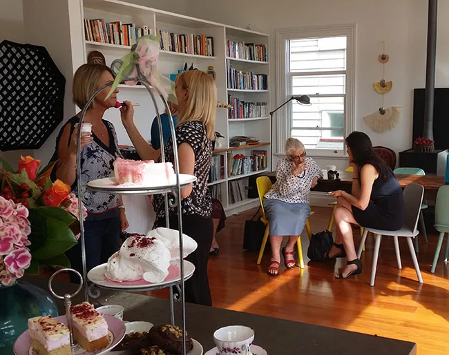 Group of people socializing in a living room with cakes and bookshelves in the background.