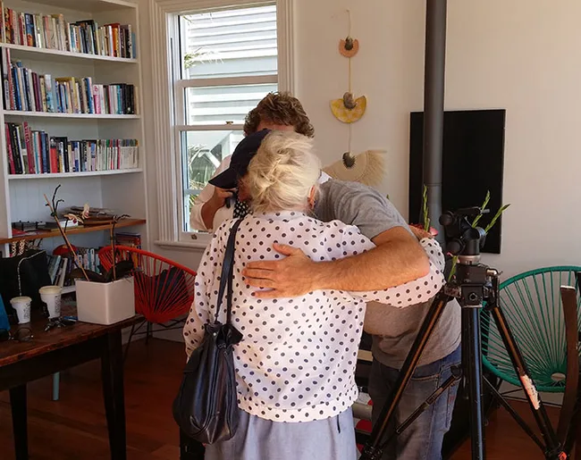 Two people embracing in a cozy living room with bookshelves and a tripod nearby.
