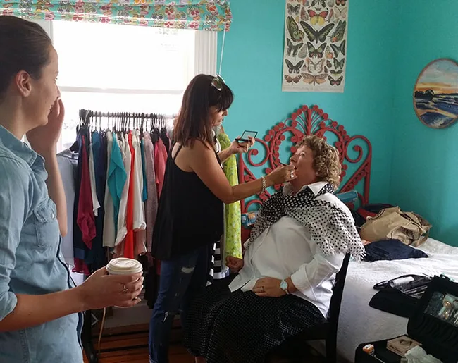 Makeup artist applying lipstick to a seated woman in a colorful room, while another woman holds a coffee cup nearby.
