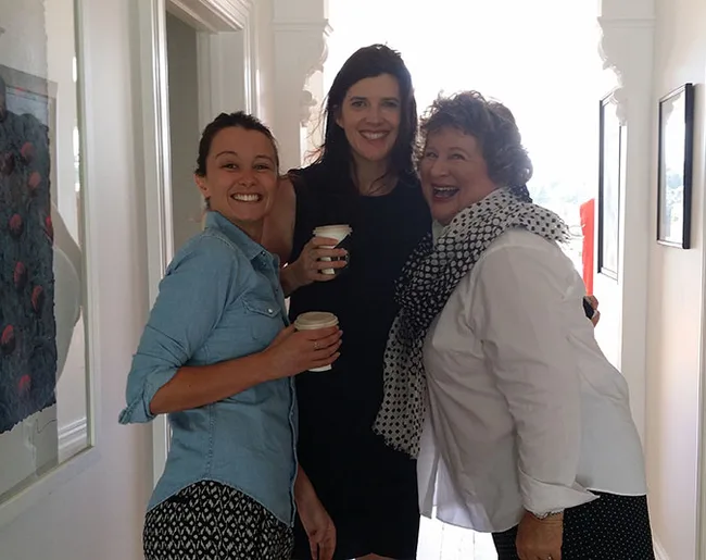 Three women smiling and holding coffee cups in a bright hallway.
