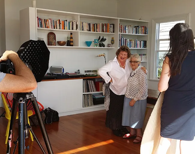 Two women posing together for a photo in a well-lit room with shelves of books in the background.