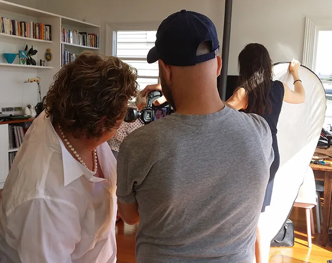 Photography crew setting up; a man adjusts camera while two women assist with positioning and lighting.