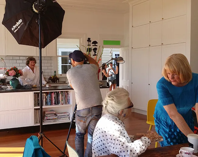 Photographer and crew taking photos in a bright kitchen with a smiling woman at the counter.