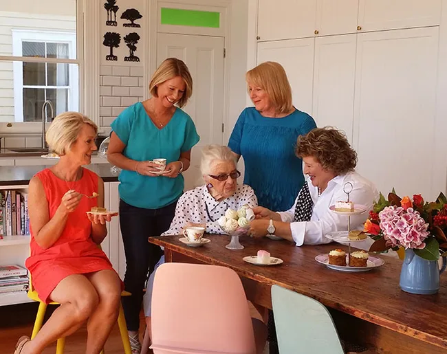 Five women enjoying tea and cupcakes around a wooden table in a bright kitchen setting.