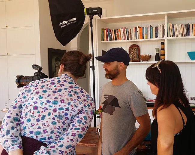 Three people stand around a camera on a tripod in a room with bookshelves and lighting equipment.