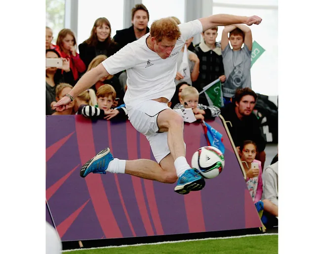 Man in white athletic wear kicking a soccer ball mid-air indoors, with spectators in the background.