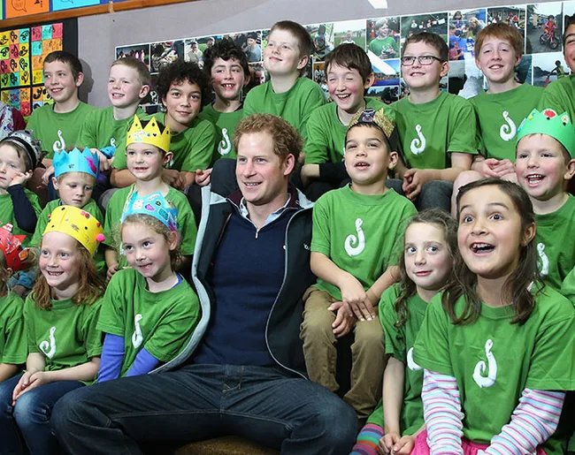A group of children in green shirts and paper crowns pose with an adult man in a casual jacket, smiling in a classroom.