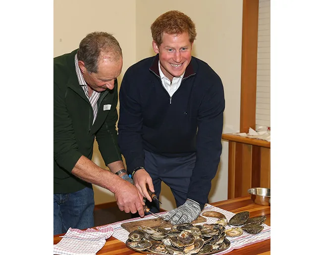 Two men opening oysters on a table, smiling, one wearing a glove and dark sweater.