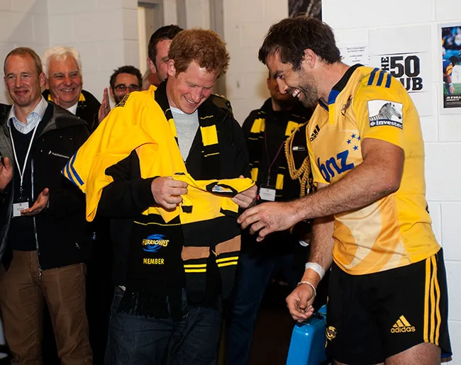 Two men laughing, one holding a yellow sports jersey. People in background clapping and smiling.