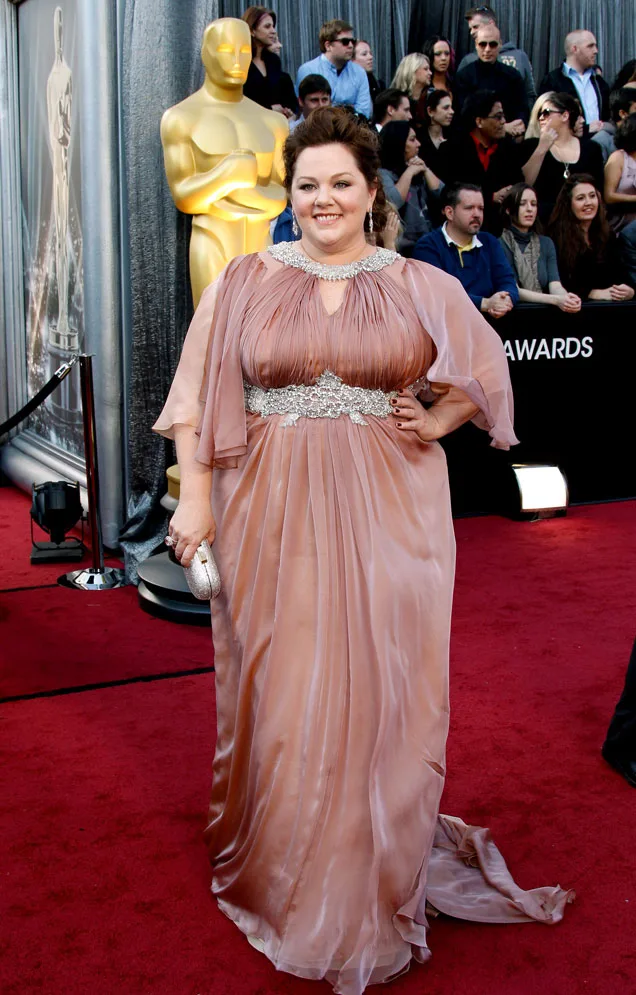 A woman in an elegant pink gown poses on the red carpet near a large Oscar statue, with a crowd in the background.