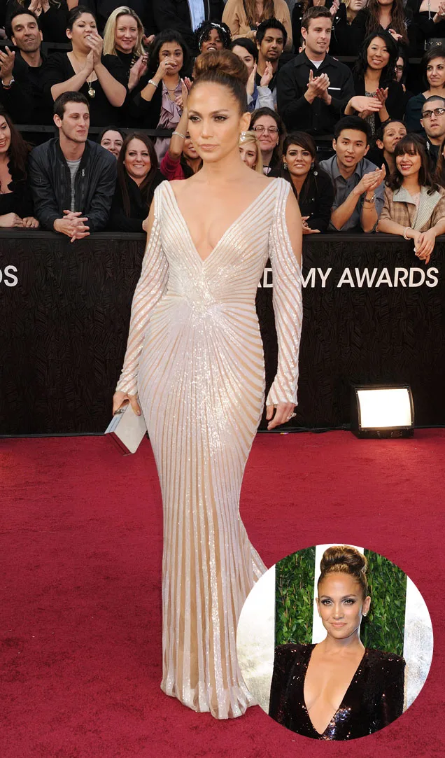 Woman in a shimmering gown on the red carpet, with a bun hairstyle, surrounded by a cheering crowd.