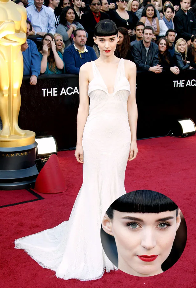 Red carpet event with individual in a white gown, crowd and large award statue in the background.