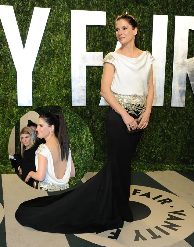 Woman in a white top and black gown with gold details, posing on a red carpet with a "Vanity Fair" backdrop.