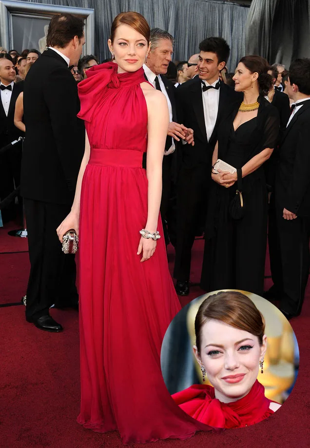 Red carpet event, woman in a red gown with a bow at the neck, elegant hairstyle, and jewelry; crowd in the background.