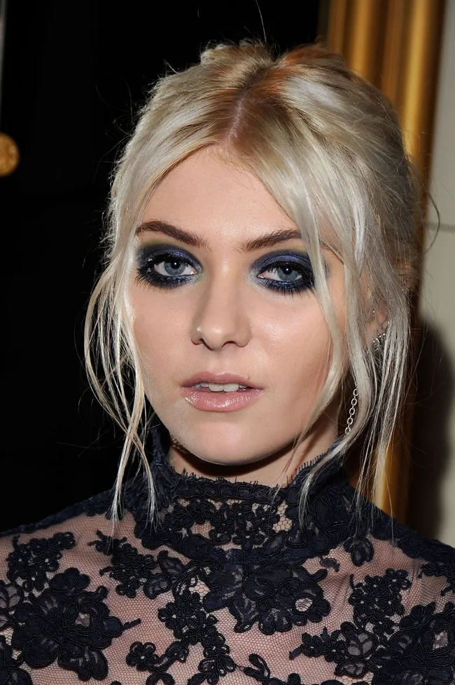 Blonde woman with blue eye makeup and lace top, looking at the camera.