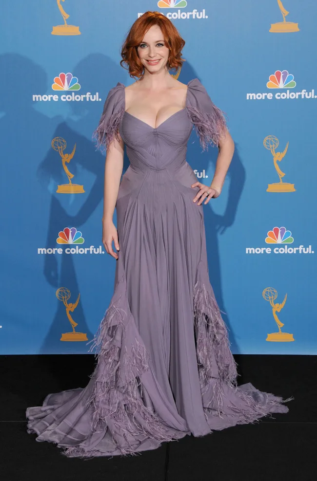 Red-haired woman in a purple gown with feather details, smiling at an awards event with a blue background.