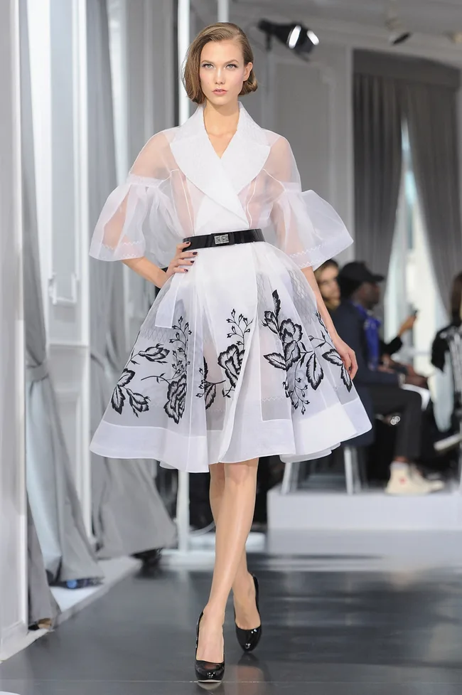 Model in a sheer white dress with black floral details walks the runway in a fashion show setting.