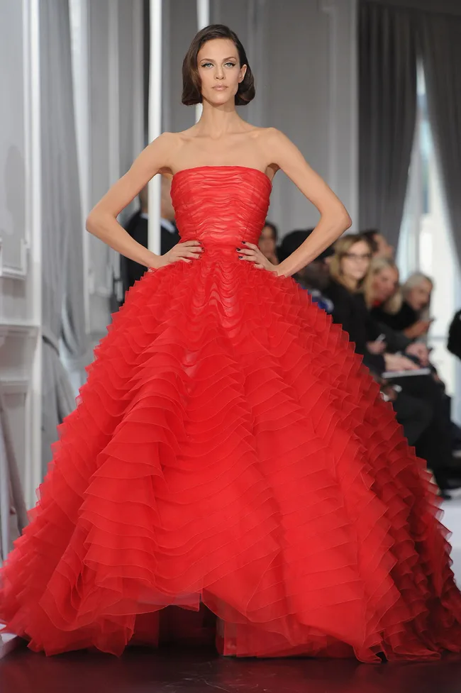 Model in an elegant, strapless red gown with ruffled layers, hands on hips, walking down a runway.