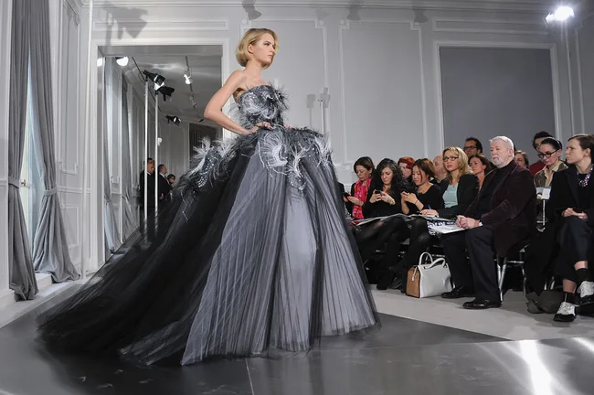 Model in a flowing gray and black gown walks the runway, watched by an audience in a fashion show setting.