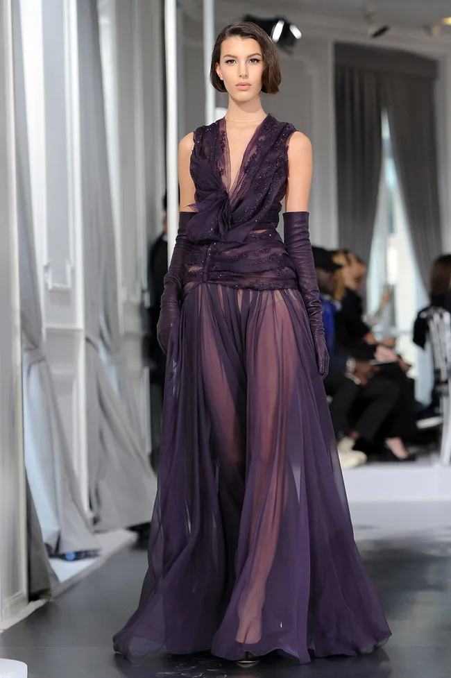 Model wearing a sheer purple gown and gloves on a fashion runway.