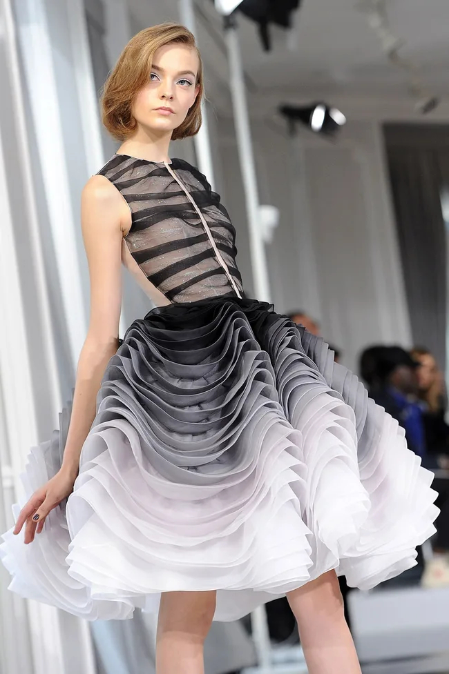 Model on runway wearing a sleeveless, ruffled gray ombr&eacute; dress with sheer black striped top, at a fashion show.