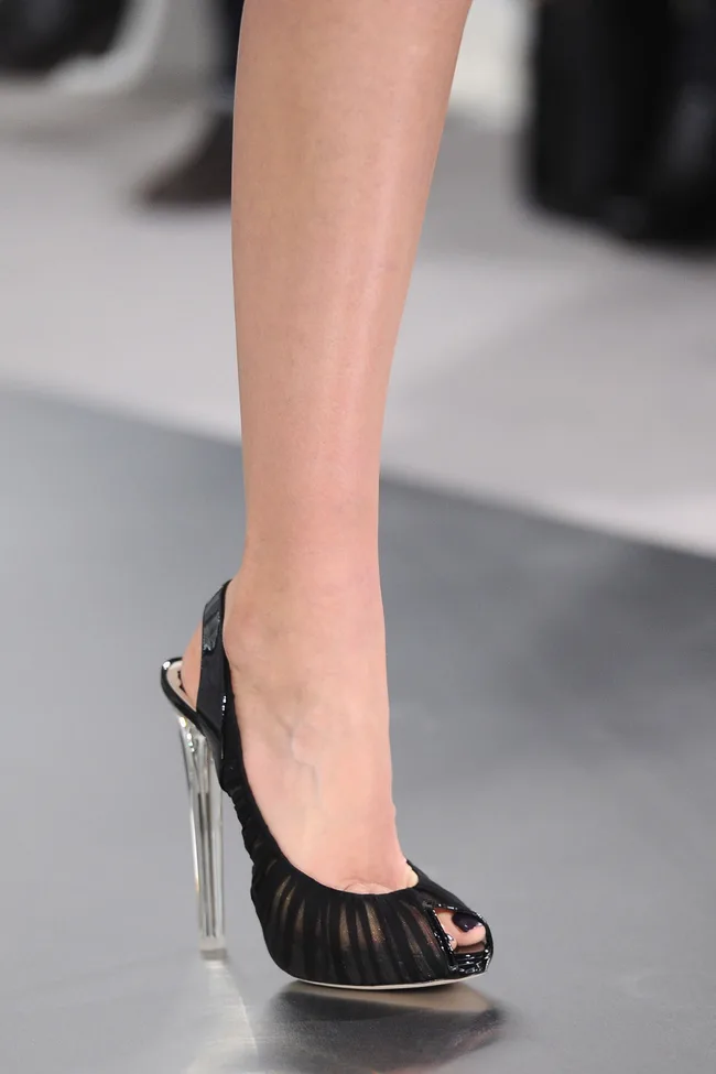 Black high-heeled sandal with transparent heel, worn on a runway.