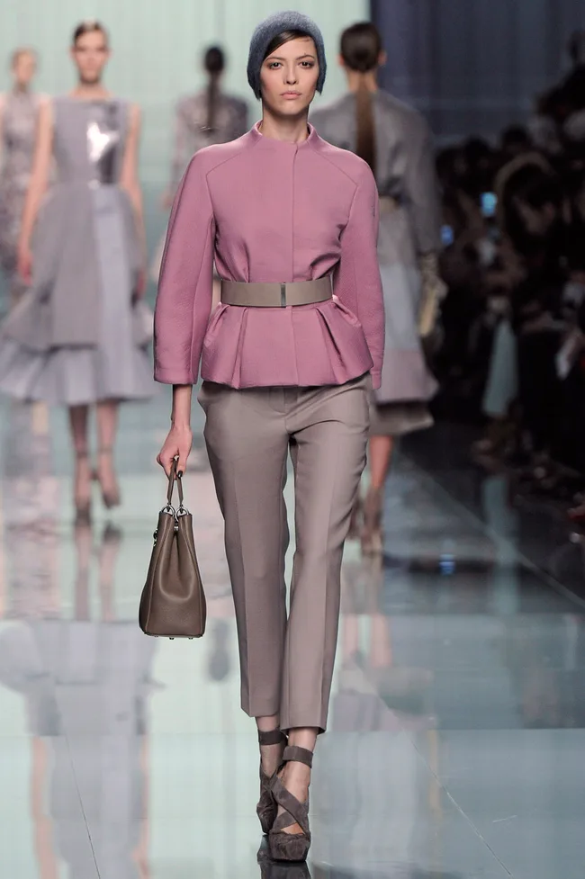 Model in a pink jacket and gray pants, carrying a brown bag, walks runway; other models in the background.