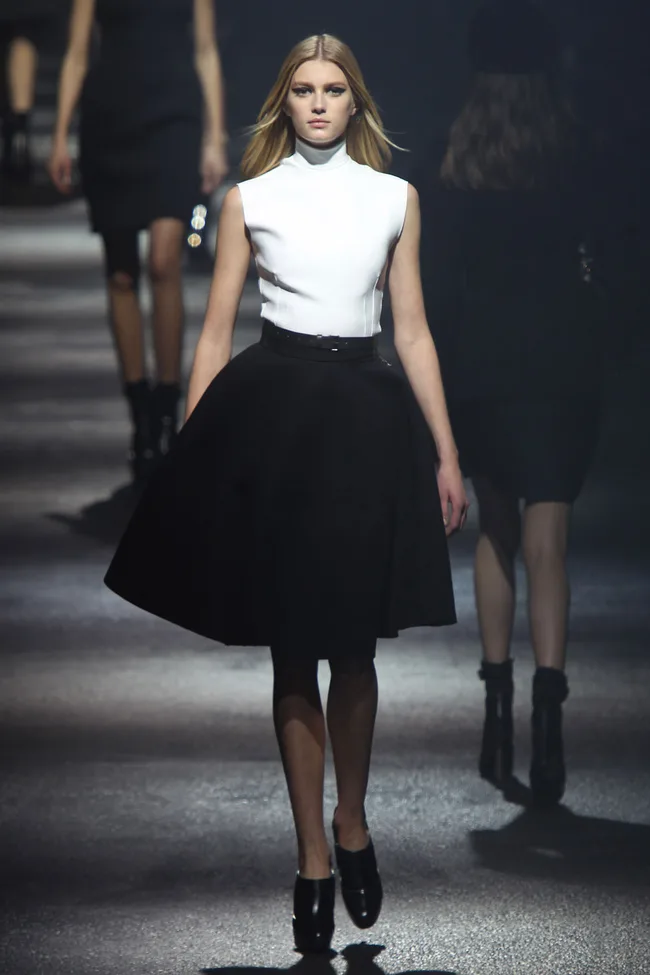 Model wearing a white sleeveless top and black skirt walks the runway in high-heeled shoes.