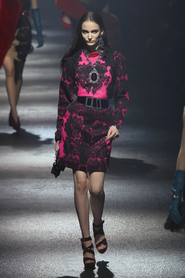 Model in a pink and black dress with bold patterns, walking a dimly lit runway wearing chunky jewelry and strappy heels.