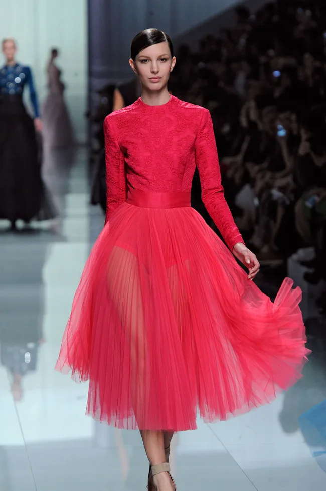 Model walking runway in a red, long-sleeve, pleated dress at a fashion show.