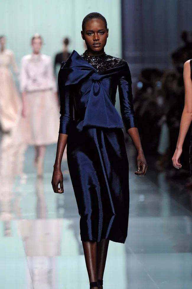 Model wearing a dark blue satin dress with a large bow walks down a fashion runway.