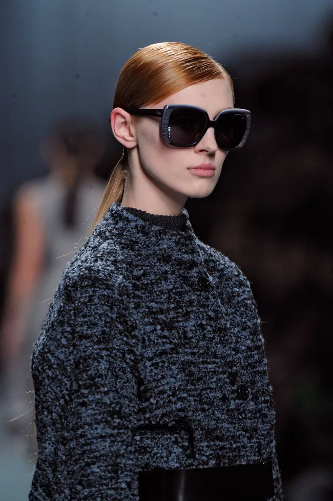 Model with slicked-back hair, wearing large sunglasses and a textured coat, walking on a dimly lit runway.