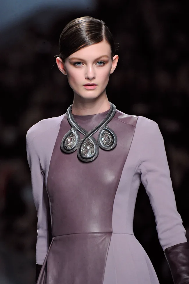 Model in a sleek, mauve dress with a statement teardrop necklace, walking the runway at a fashion show.