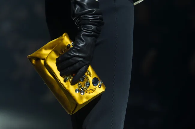 A person wears black gloves and holds a yellow clutch adorned with jewels.