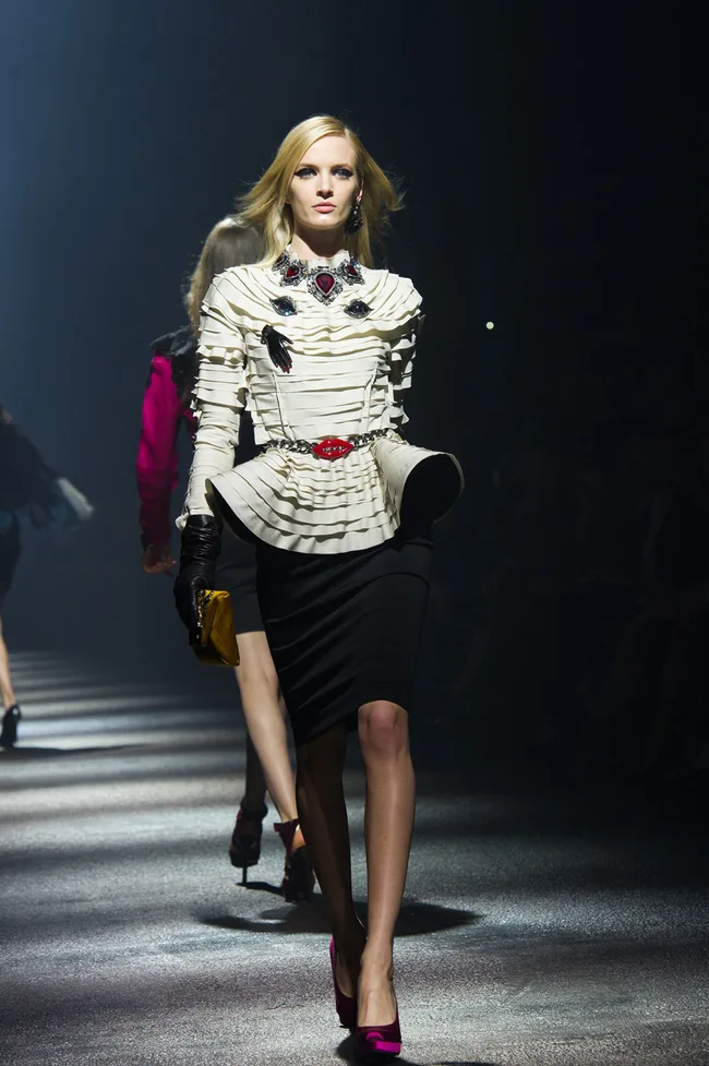 Model on runway in white ruffled top, black skirt, holding a yellow clutch, with dramatic lighting.