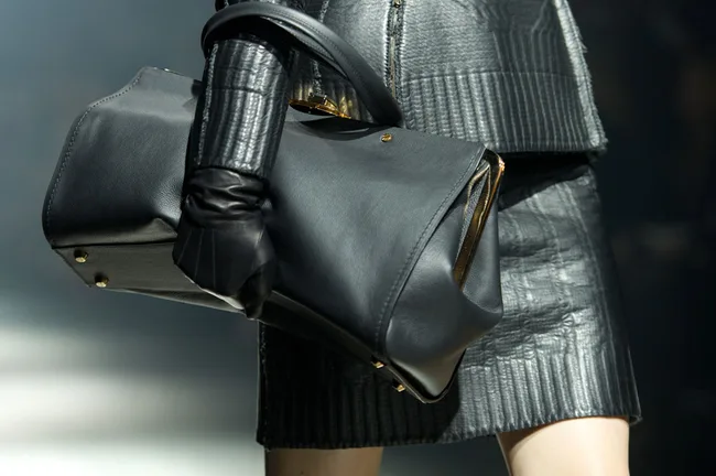 A model in black leather outfit holds a large black leather handbag with gold accents on the runway.