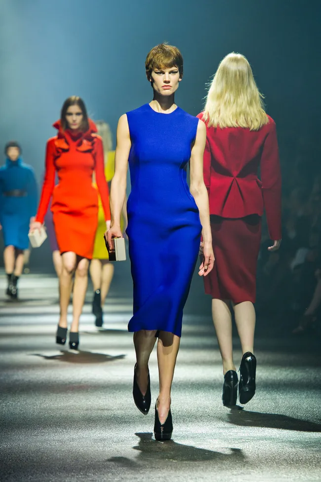 Models on a runway, the foremost in a blue dress, with others behind in colorful outfits.