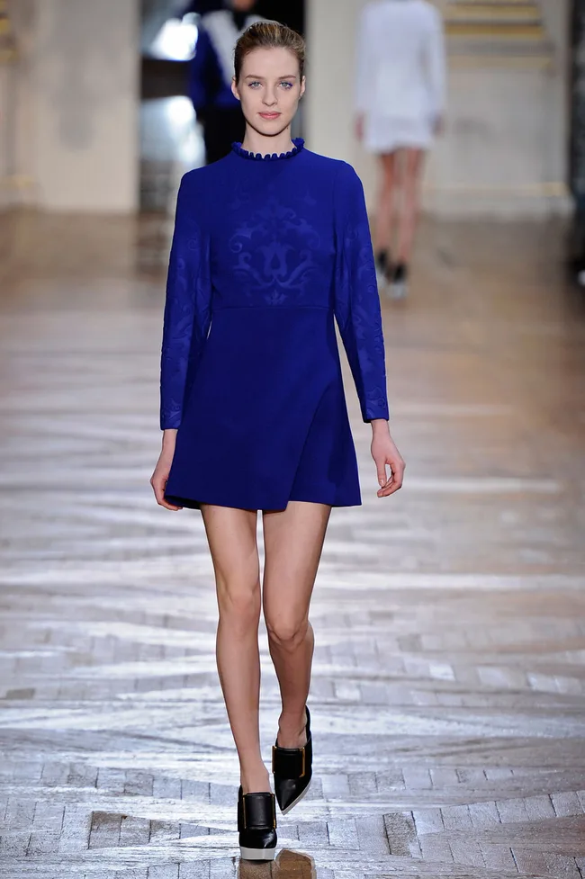 Model in a blue patterned dress with black shoes walking on a runway.