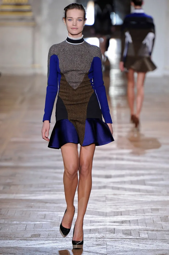 Model walking on a runway wearing a multicolored dress with a flared hem, featuring shades of brown, black, and blue.