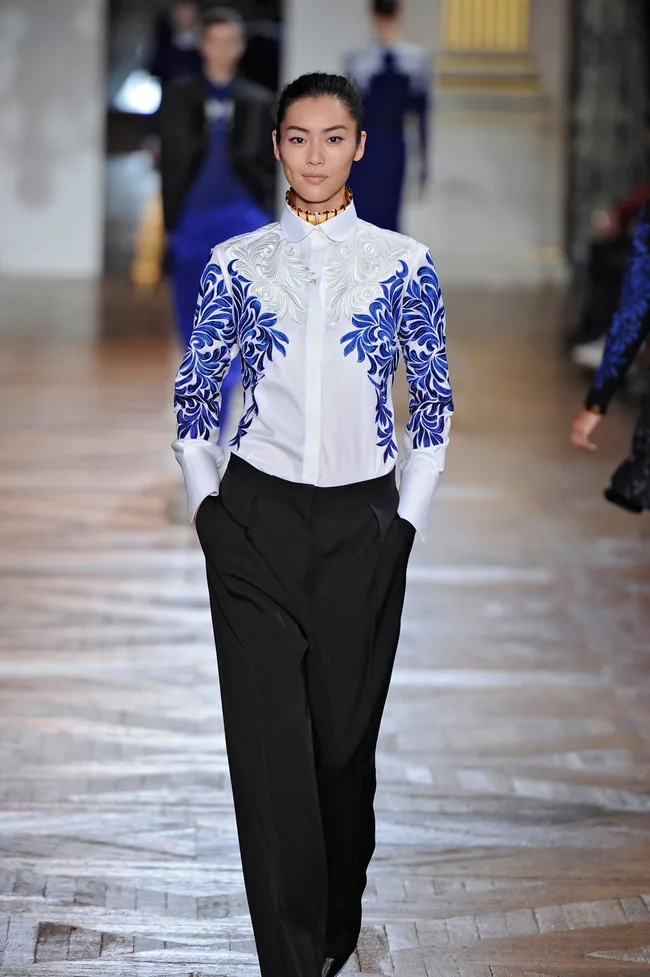 Model walking in a fashion show wearing a white shirt with blue floral designs and black pants.