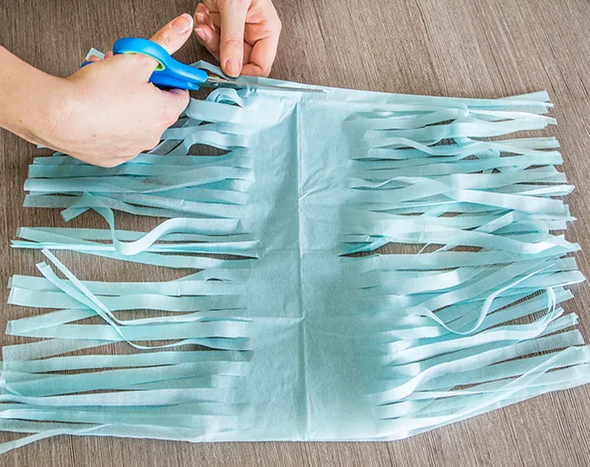 Hands using scissors to cut blue tissue paper into fringes on a table.