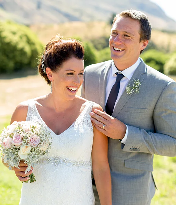 Bride and groom smiling outdoors; bride in white dress holding bouquet, groom in gray suit with tie.