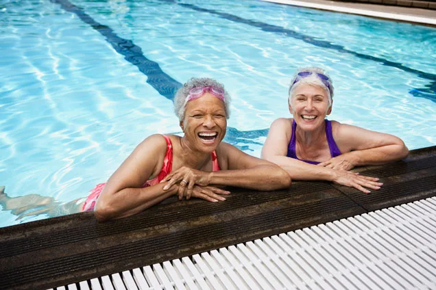 Two elderly women smiling and relaxing by the edge of a swimming pool, wearing swimsuits and swim caps.