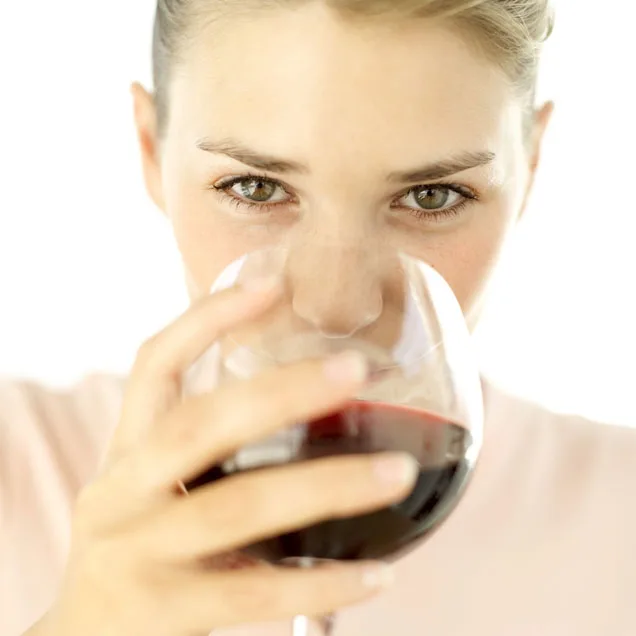 A woman with light hair holds and drinks from a glass of red wine, gazing directly at the camera.